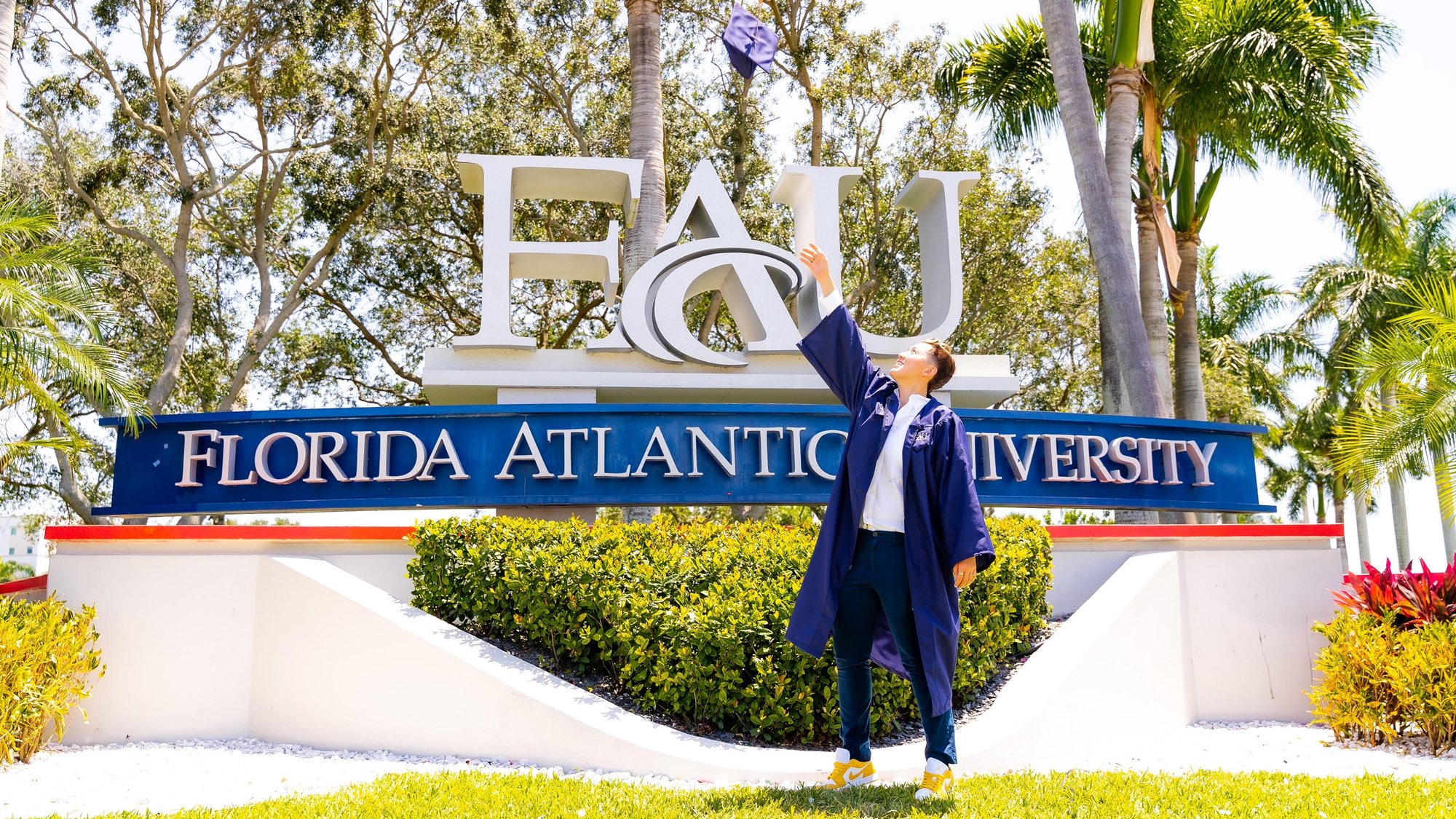 Ashley throwing her cap into the air in front of the FAU school sign upon graduation.