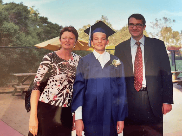 Alistair, in his cap and gown, in between his parents at 8th Grade graduation.